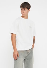 Witte katoenen t-shirt met een ronde hals en korte mouwen. Bevat een klein grijs embleem op de linkerborst. Gecombineerd met lichtgrijze jeans.