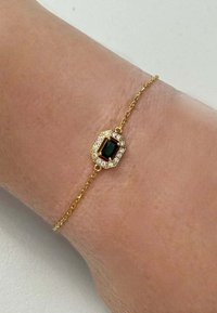 Bracciale in oro con al centro una pietra verde ottagonale, circondata da piccole pietre trasparenti, su un polso in primo piano su uno sfondo neutro.