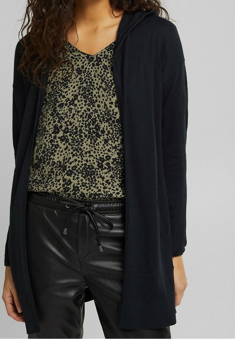 Black hooded cardigan over a green top with black floral pattern, paired with black leather pants featuring an adjustable drawstring waist.