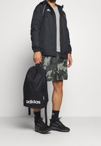 Černá dešťová bunda s bílými detaily, šedé tričko, maskáčové šortky, černý batoh Adidas s bílým logem a černé sportovní boty.