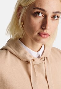Sweatshirt à capuche beige en tissu texturé, avec un cordon de serrage, porté sur une chemise blanche à col. Focus sur le visage et le haut du torse.