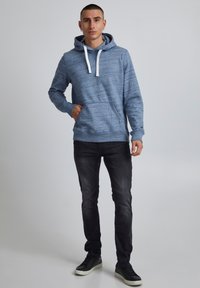 Ljusblå huvtröja med ett strukturerat mönster, vita dragsnören och kenguruficka, i kombination med åtsittande svarta jeans och svarta sneakers.