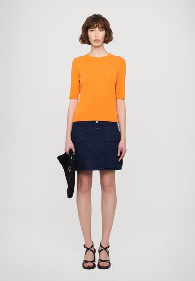 Oranžna rebrasta majica s kratkimi rokavi, kombinirana z mornarsko modrim denim krilom z velikimi žepki, dopolnjena z črno tkaninasto torbo in sandali z naramnicami.