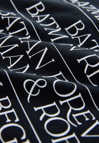 Tela negra con texto blanco repetido que incluye nombres y símbolos. El material parece suave, con una textura lisa y pliegues ligeramente estirados.