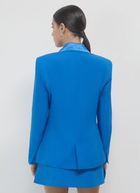 Blazer blu dal taglio slim, texture liscia, rever a lancia e spacco singolo sul retro. Abbinato a una gonna coordinata.