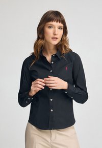 Maglietta nera a maniche lunghe con collo a camicia, bottoni bianchi e un piccolo logo rosso sul petto; tessuto morbido con una vestibilità sartoriale.