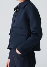 Giacca blu navy realizzata in tessuto liscio, dotata di colletto, chiusura frontale con zip e due tasche a toppa sul petto.