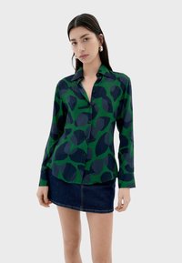Camisa de botones en verde y azul marino con un patrón de hojas; mangas largas; cuello; combinada con una minifalda de mezclilla. Textura de tela suave.