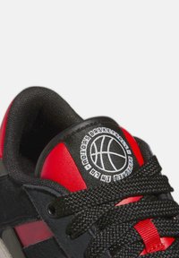 Baskets de basket-ball noires et rouges en matériaux synthétiques et en daim lisses, avec des lacets texturés et un logo circulaire représentant un ballon de basket.