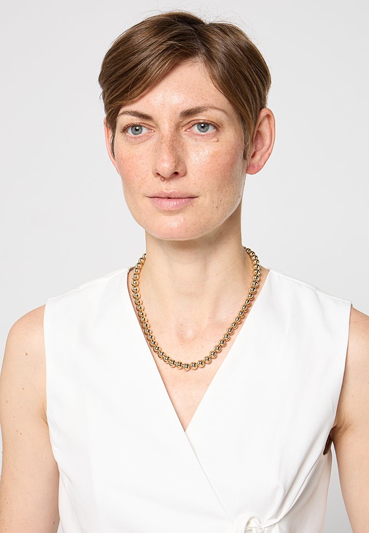 Collana in metallo dorato con perline rotonde, indossata su un top bianco senza maniche, con design a drappeggio e un dettaglio annodato in vita.