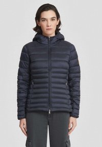 Vrouw in een zwarte gewatteerde jacket met capuchon en rits aan de voorkant, staand tegen een effen lichte achtergrond, met het gezicht naar voren en een neutrale uitdrukking.