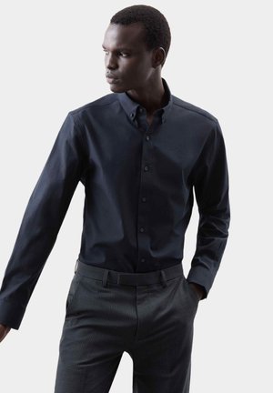 Granatowa koszula z długim rękawem, zapinana na guziki, z kołnierzykiem, w połączeniu z wzorzystymi, ciemnoszarymi spodniami. Gładki materiał o dopasowanym kroju.