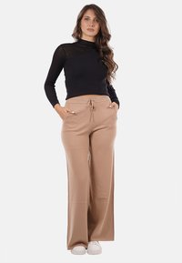 Pantaloni beige a gamba larga con vita regolabile, abbinati a un top nero a maniche lunghe a coste; il modello sta in piedi con le mani in tasca.