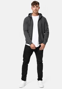 Haut de sweat à capuche gris avec cordons noirs, porté par-dessus un T-shirt blanc, associé à un jean noir et des baskets blanches. Le modèle se tient droit, les mains visibles.