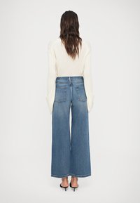 Mulher com cabelo castanho longo veste um suéter creme em malha canelada, jeans azul de cintura alta e pernas largas, e sapatos de salto abertos pretos, de pé contra um fundo branco.