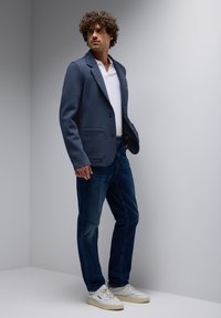 Dunkelblazer mit Reverskragen, weißem Poloshirt, dunklen Jeans und weißen Sneakers. Glatte Textur und taillierte Passform.