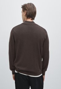 Maglione marrone a maglia con collare e polsini a coste, maniche lunghe e vestibilità rilassata, indossato sopra una camicia bianca. Texture liscia.