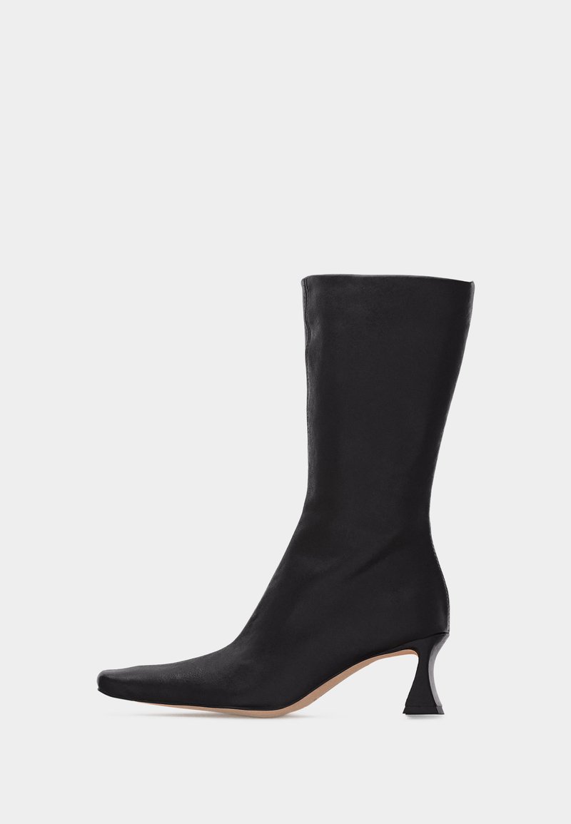 Μαύρη δερμάτινη μπότα μέχρι τη μέση της γάμπας με μυτερή μύτη και καμπυλωτό χαμηλό τακούνι σε απλό ανοιχτό φόντο.