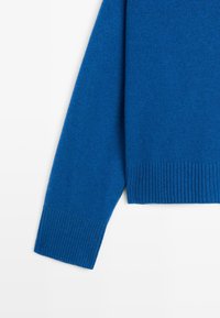 Manica di maglione blu lavorato a maglia e orlo inferiore con polsini a coste su sfondo bianco.