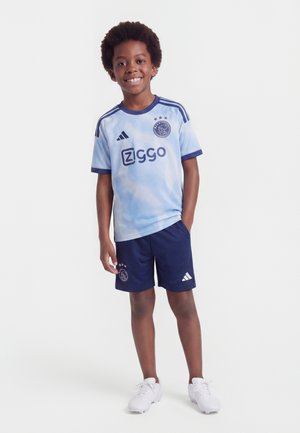 Niño sonriendo, vestido con uniforme de fútbol azul claro y azul marino con el logo de Ziggo, de pie con una mano en el bolsillo y zapatillas blancas sobre fondo liso.