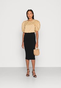 Blusa com mangas bufantes em mostarda com padrão floral branco, combinada com uma saia midi preta canelada e uma pequena bolsa de palha; sandálias de salto pretas.