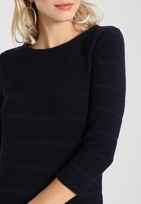 Top a rayas marino con mangas tres cuartos, que presenta una textura suave y un escote redondo. Grandes pendientes de aro complementan el diseño.