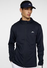 Felpa con zip di colore navy realizzata in materiale morbido e elasticizzato, con cappuccio e due tasche laterali. Un logo bianco è visibile sul petto.