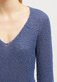 Nahaufnahme einer Person, die ein blaues, strukturiertes Strickoberteil mit V-Ausschnitt und langen Ärmeln vor einem einfarbigen hellen Hintergrund trägt.