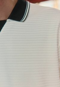 Bílý polo tričko s texturou a černým límečkem, který je ozdoben dvěma tenkými bílými pruhy. Tkanina má vodorovné žebrování.