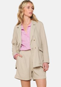 Beige Leinen-Doppelreiher-Blazer über einem pinken Hemd mit Knöpfen, kombiniert mit beigen Shorts. Glatte Textur, strukturiertes Design und sichtbare Taschen.