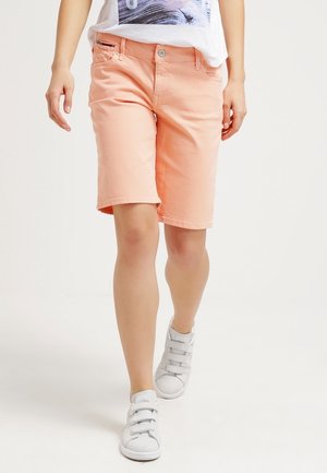 Person iført ferskenfarvede knælange shorts, en hvid t-shirt med grafik og hvide sneakers, stående mod en ensfarvet lys baggrund.