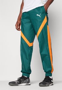 Pantalon de jogging vert avec des bandes diagonales orange, poignets élastiques, poches latérales et logo Puma blanc. Fabriqué à partir d'un matériau lisse et léger.