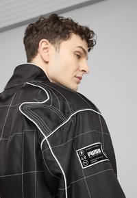 Jachet�ă de curse neagră cu cusături albe, având un guler structurat, un patch cu marcă și un model în rețea pe un material durabil.