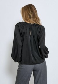 Schwarze Satinbluse mit rundem Ausschnitt, gerafftem Rückendetail und Puffärmeln, kombiniert mit grauen, taillierten Hosen.