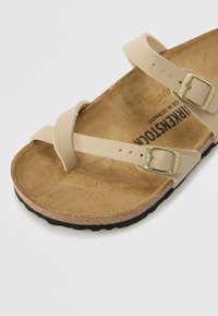 Beige nahkasaapad, millel on kaks rihma ja kuldsed pandlad, korkjalats ja must kummitald, millel on sile tekstuur ja lihtne disain.