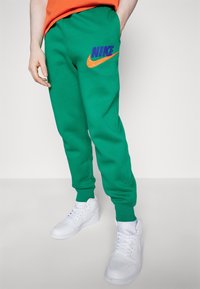 Zelené tepláky Nike s oranžovo-fialovým vyšívaným logem, elastick ými manžetami a bočními kapsami, doplněné o bílé tenisky.