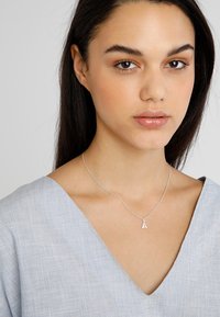 Collana d'argento con pendente a forma di lettera "A", caratterizzata da una catena sottile. Indossata su una blusa leggera di colore blu chiaro, con scollatura ampia.