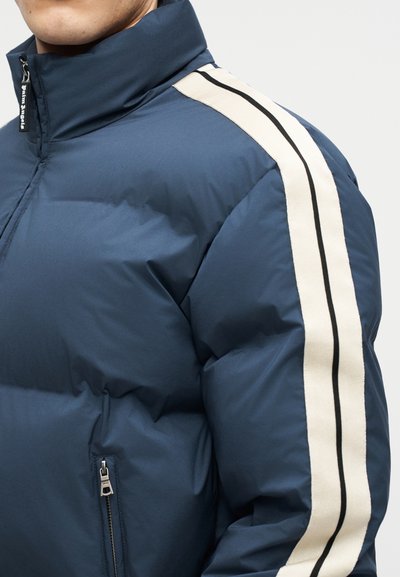 Tumesinine sulejope kõrge kraega, valge õlanöör musta triibuga ja lukuga küljetaskuga, mida kannab inimene, keda näidatakse kaelast allapoole.