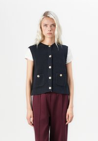 Marine gebreide vest met ronde hals, knopen aan de voorkant en twee voorzakken. Draag het over een wit shirt, gecombineerd met bordeauxrode broeken.
