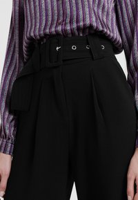 Svarta högmidjade byxor med veck, med ett brett bälte med silverfärgade öglor. Matchas med en knäppt skjorta med lila ränder.