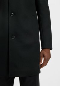 Cappotto nero in lana con due bottoni, revers a scollo a V e tasca laterale. Il tessuto ha una texture liscia e una vestibilità sartoriale.
