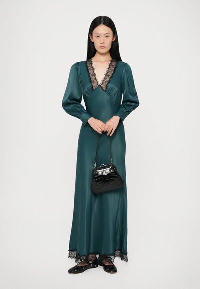 Lang, smaragdgroene satijnen jurk met een diepe, kantafgezette decolleté en lange mouwen, in combinatie met een zwarte croc-geembossede handtas en platte schoenen.