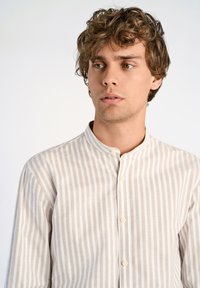 Camicia bianca con sottili strisce verticali beige, tessuto leggero, colletto a fascia e chiusura frontale con bottoni. La superficie strutturata aggiunge un dettaglio sottile.