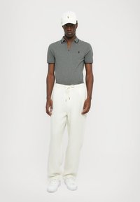 Cămașă polo gri cu mâneci scurte, logo alb și pantaloni de culoare crem. Adidașii albaștri completează ținuta. Materialul pare moale și lejer.