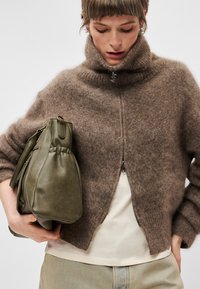 Sweater castanho felpudo com fecho éclair, colarinho alto e punhos canelados, usado sobre uma camisa clara, acompanhado de uma mala de couro verde.