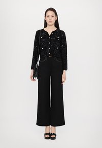 Cardigan cu mărgele negre, cu nasturi, asortat cu pantaloni largi negri cu talie înaltă. Modelul poartă o geantă mică din piele cu franjuri negri și sandale negre cu platformă.