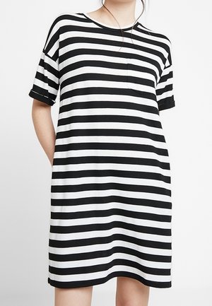 Personne portant une robe ample mi-genoux avec des rayures horizontales noires et blanches et une petite poche poitrine, debout avec une main dans la poche.