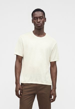 Ung mand iført en ensfarvet, off-white kortærmet skjorte og brune bukser, stående mod en ensfarvet lys baggrund.