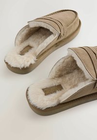 Hnedé semišové papuče s plyšovým krémovým kožúškom, ktorý má uzavretý dizajn a jemné dekoračné švy. Protišmykové podrážky.