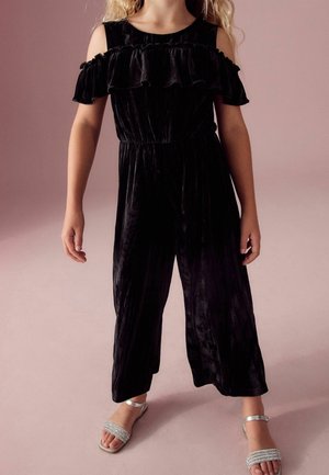 Combinaison en velours noir avec design décolleté, col à volants et rayures verticales ; associée à des sandales argentées scintillantes.